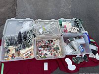Three clear plastic storage bins filled with assorted Christmas village accessories on red cloth
