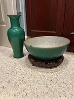 Photo showing the green enameled vase and large bowl on a carpeted floor with wooden doors in background. The bowl rests on a carved wooden stand.