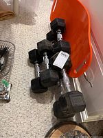 Photo showing the 3 pairs of dumbbells arranged on carpet near an orange plastic item and a white cabinet. We see the rubber ends marked 10, 15, and 20 lbs.