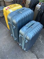 Three hard shell luggage pieces: two blue Eminent suitcases in front and one large yellow TravelPro suitcase behind, all upright on asphalt ground.