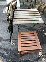 Photo showing the square wood slatted table with two matching folding chairs propped against it and smaller teak side table in front.