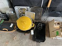 Large yellow corn pot with lid atop black electric grill, surrounded by aluminum roasting and baking pans, and wire racks on concrete floor.