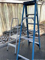 Photo showing both ladders side by side outdoors on a paved surface with a wooden fence and gardening stones in the background.