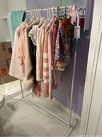 Wide angle view of the white metal portable clothing rack holding several ladies clothing items including sweaters, blouses, and skirts in pastel colours.