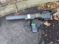 Full view of WORX 20V cordless leaf blower laid on the ground with attached nozzle, black and orange colors visible. Battery charger shown next to blower with power cord.