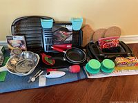View of lot including BBQ grill pan, steamer basket, frying pans, glass storage containers with lids, cork trivets, plastic wrap, and kitchen tools