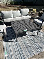 Square metal slat coffee table placed outdoors on a patterned rug between patio seating, showing overall shape and condition.