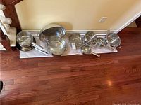 Full view of all cookware lined against the wall on floor including pots, pans, lids and wok
