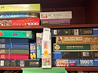 Shelf with large collection of jigsaw puzzles stacked, including boxes from Ravensburger, Cobble Hill, Falcon, and others, showing multiple 500 and 1000 piece puzzles with various themes.
