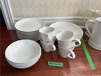 Photo showing stacks of white dinner plates, luncheon plates, bowls, and grouped mugs on wooden floor with green tape and Royal Doulton label.