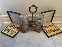 Full lot view showing two spoon sets in cases flanking ornate silver-plated condiment stand with cut glass jars