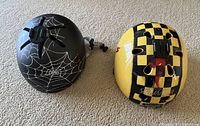 Top view of two children's bike helmets showing external designs: specialized yellow and black checkered helmet and black Nutcase helmet with white spider web.