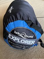 Photo showing black and blue Lucky Bums Explorer mummy sleeping bag in compression sack with adjustable shoulder strap.