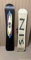 Photo showing bottoms of the two snowboards: black K2 brand board and a heavily worn black and white board with large block letters.