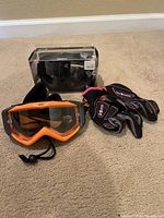 Photo shows two pairs of clear motocross goggles placed side by side. One has an orange frame with a black tiger stripe pattern strap and the other has a black frame with black strap. Also includes a pair of black motocross gloves with pink detailing, size 7.