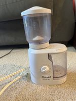 White DeLonghi electric coffee mill with clear plastic bean hopper and grounds container visible. Adjustable grind dial and power cord shown.