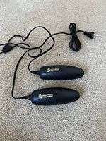 Pair of black electric boot/shoe dryers or heaters with attached power cords and standard plugs laid out on carpet.