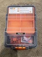 Top view of the closed storage case showing the orange latches, clear plastic lid with compartments, and Flexo label with warnings and suitability info.