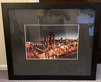 Front view of the framed night cityscape photograph showing steel bridge structure and city lights, black matte and frame visible.