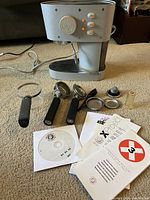 Francis Francis X3 espresso machine with three portafilter handles, filter baskets, tamper, and manuals laid out on carpet.