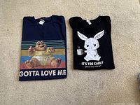 Two folded graphic T-shirts on carpet. Left is navy blue 'Gotta Love Me', right is black bunny 'It's Too Early'.