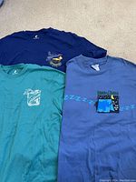 Three T-shirts laid on carpet floor, showing front graphics of navy, green, and blue shirts.