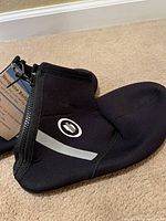 Black neoprene booties with zipper and reflective strip, close-up side view showing logo and material texture.