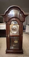 Front view of Sligh mahogany wall clock showing ornate dial, moonphase dial, brass pendulum behind glass door, and detailed wood casing with arch top.