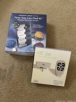 Photo of two boxed small electronic devices on carpet: a wireless electronic locator with colorful key fobs and an ABT iJet RF remote for iPod.