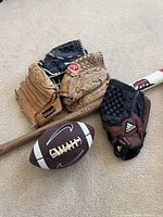 Four baseball gloves of various sizes, a Nike football in front, and part of a wooden baseball bat with a red and white handle and bear graphic.