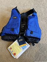Front side view of blue NRS youth life preserver with tags attached, showing pockets and adjustable straps.