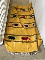 Full view of the yellow woven blanket with colorful bird motifs and fringe edges laid flat on carpet.