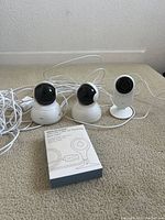 Three Yi home security cameras placed on carpet with white USB cables connected, alongside a USB power adapter box for charging.