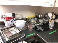 Wide view of assortment of bakeware and kitchen items on countertop showing metal baking sheets, mixing bowls, a Pyrex measuring cup, Sunbeam hand mixer box, kitchen utensils, and towels.