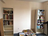 Front view of the two white wooden bookshelves against a wall. One bookshelf contains books; the other has puzzles and games. Both show signs of wear.