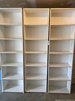 Photo shows three tall white bookshelves standing side by side in a garage, each with five fixed shelves, showing wear and damage.