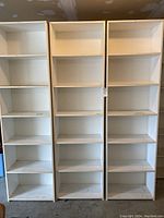 Photo showing three tall white bookshelves side by side against a garage wall, each with multiple fixed shelves and visible wear and damage.