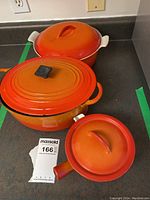 Three orange ombré cast iron enamel dishes with lids on countertop, includes small sauce dish, round casserole, and oval casserole dishes