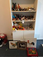 Shelf displaying multiple chicken and rooster figurines made from wood, ceramic, and glass along with plush toys and painted panels.