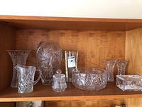 Photo showing assortment of crystal and glass items on wooden shelf, including vases, serving bowl, sugar dish, small pitcher, and glass holder.