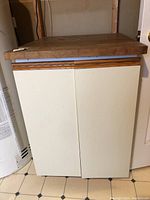 Full view of two-door cupboard with cream-colored doors and wooden removable top, placed on tiled floor against wall.