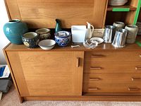 Photo showing all items in the lot arranged on a wood sideboard including pottery, planters, pewter tankards, steins, white pitcher, and glass ashtray.