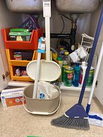 Front view of cleaning supplies stored under sink including broom, mop bucket, orange plastic organizer, and various cleaning products