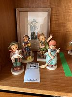 Four Hummel figurines arranged on shelf with framed lithograph print behind them.