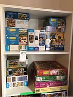 Shelves displaying multiple stacks of jigsaw puzzle boxes from brands including Charles Wysocki, MB, Hometown Collection, and others.