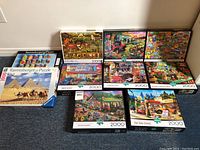 Photo showing 11 jigsaw puzzle boxes including Buffalo and Ravensburger brands, featuring 1000 and 2000 piece counts and various scenic images: pyramids, outdoor markets, post office, curiosity shop with cat.