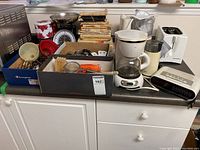 Overview of kitchen lot showing coffee maker, toaster, food processor, kitchen scale, alarm clock, and boxes of utensils and miscellaneous items on white counter.