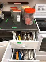Plastic food storage containers with orange and beige lids on countertop. Two drawers open showing various everyday cutlery including forks, spoons, and knives with plastic handles.