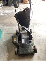Front view of the electric lawn mower with visible wheels, handle, and bag attachment.