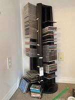 Black plastic CD tower holding approximately 30-40 CDs, standing on carpeted floor against wall, with additional CDs and cassettes stacked at base.